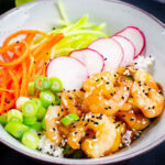 Honey and Garlic prawn poke bowl served with grated carrot,cucumber ,radishes and spring onion in a green bowl of white rice.