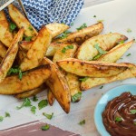 spicy curried potato wedges sprinkled with chopped basil, pouring out of a silver fry basket on white parchment paper with bbq sauce on the side