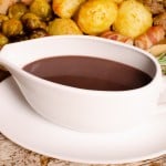 Turkey Gravy with Red Wine in a white gravy boat and saucer served next to stuffing balls,flawless roast potatoes, glazed carrots, Brussel sprouts,pigs in blankets and a roast turkey on a wooden chopping board