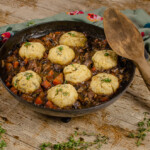 Beef and Red Wine Stew with Dumplings in a cast iron pan served on a wooden board with sprinkled sprigs of thyme, large wooden spoon on the side and a green and flowery cloth in the back ground