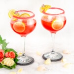 Pink Gin Fizz served in sugar rim glasses garnished with lemon slices and cubes of ice in the glass and pink and white roses at the bottom of glass