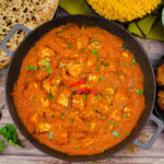 Chicken Pathia Curry in a cast iron pan with two red chillies on top and in the background there is a naan bread, pilau rice, onion bhajis and served on the side