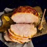 Honey Roast ham joint coated in a honey and mustard glaze on a wooden board with three slices cut from it with a honey stick coated in honey and a dollop of mustard on the sides