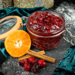 Cranberry sauce in a glass jar with a slice of clementine, two cinnamon sticks and fresh cranberries on the side served on a piece of black slate