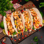 Four shrimp wraps filled with salad, king prawns and coated in sweet chilli sauce on a wooden chopping board with chopped red chillies around the tacos