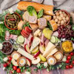 Cheese ploughman's platter served on a large wooden board which consists of crusty bread,blue cheese, cheddar cheese, brie, pork pie ,scotch egg,slices of ham, red grapes,slices of apple, cornichons, pot of pickle, pot of piccalilli and salad