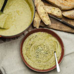 Broccoli and Stilton Soup Recipe in a bowl with stilton crumbled on top with slices of crusty bread on a board and a pot full with soup in the background