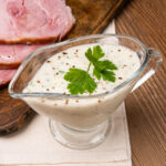Parsley Sauce Recipe in a glass sauce jug with a fresh parsley and pepper on top with slices of gammon in the background