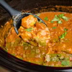 Slow cooker chicken curry with coriander leaves on top inside a black crock pot with a black spoonful of chunks of chicken in curry sauce being scooped up to serve.