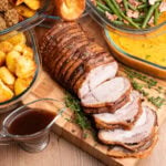 Roast Pork Loin Joint which has had three slices cut on a wooden board with roast dinner trimmings roast potatoes, sage and onion stuffing balls, honey roast parsnips, Yorkshire puddings, green beans with bacon and garlic and buttered swede and carrot mash