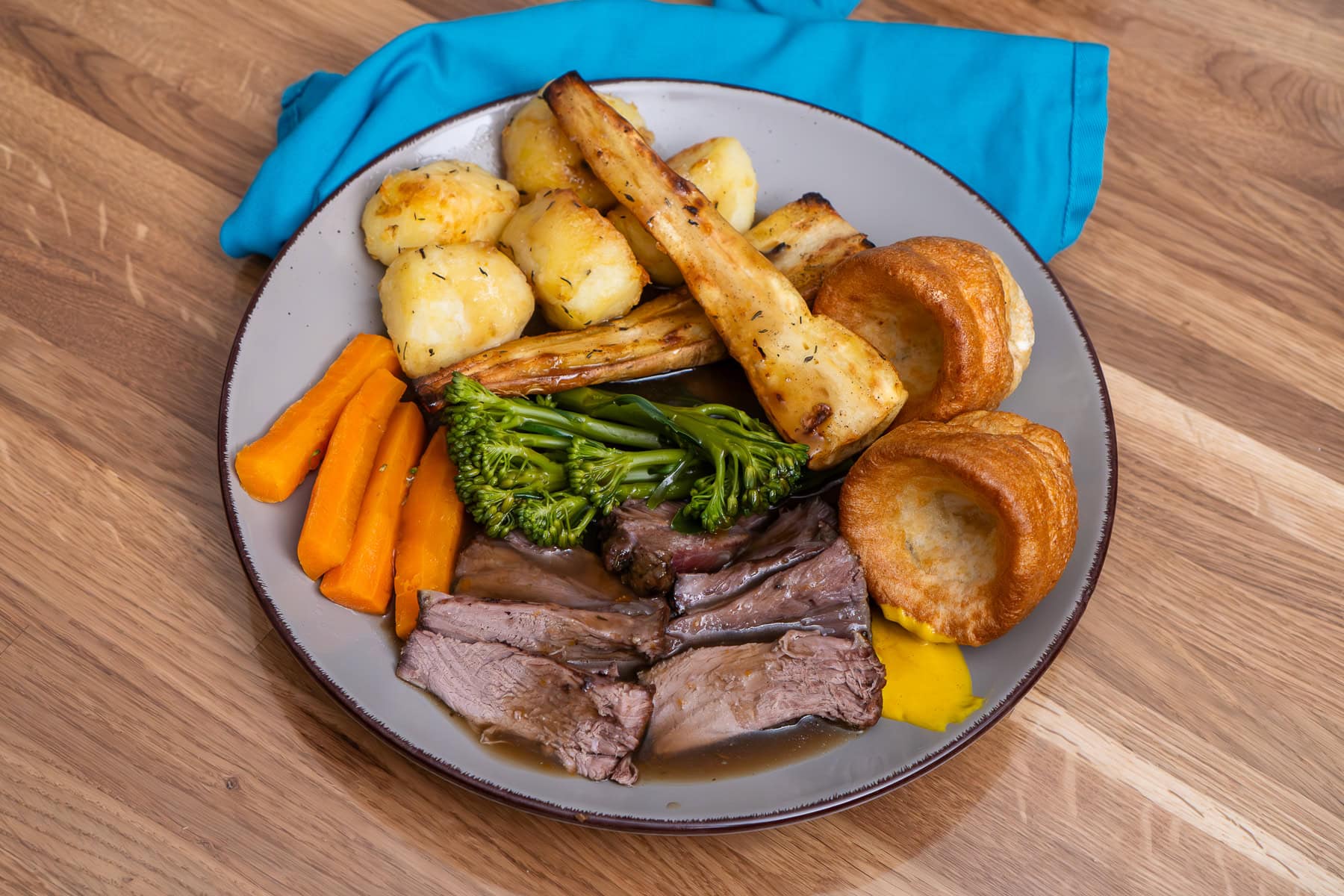 A plate of roast beef dinner which consists of slices of beef shin ,carrots, broccoli, honey roast parsnips, roast potatoes, a dollop of English mustard and Yorkshire puddings smother in gravy.