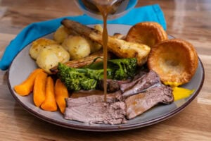 Pouring gravy over a roast dinner which consists of slices of beef shank ,carrots, broccoli, honey roast parsnips, roast potatoes, a dollop of English mustard and Yorkshire puddings.