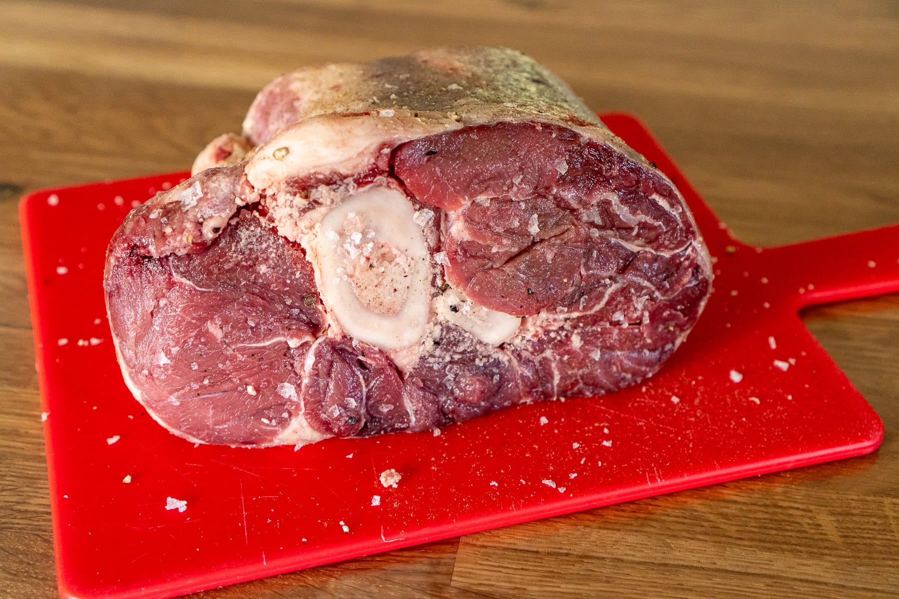 A beef shin on a red chopping board coated in salt and pepper