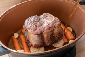 Pouring beef stock into a slow cooker pot with a beef shin sitting on top a veg trivet which consists of onions carrots and garlic cloves