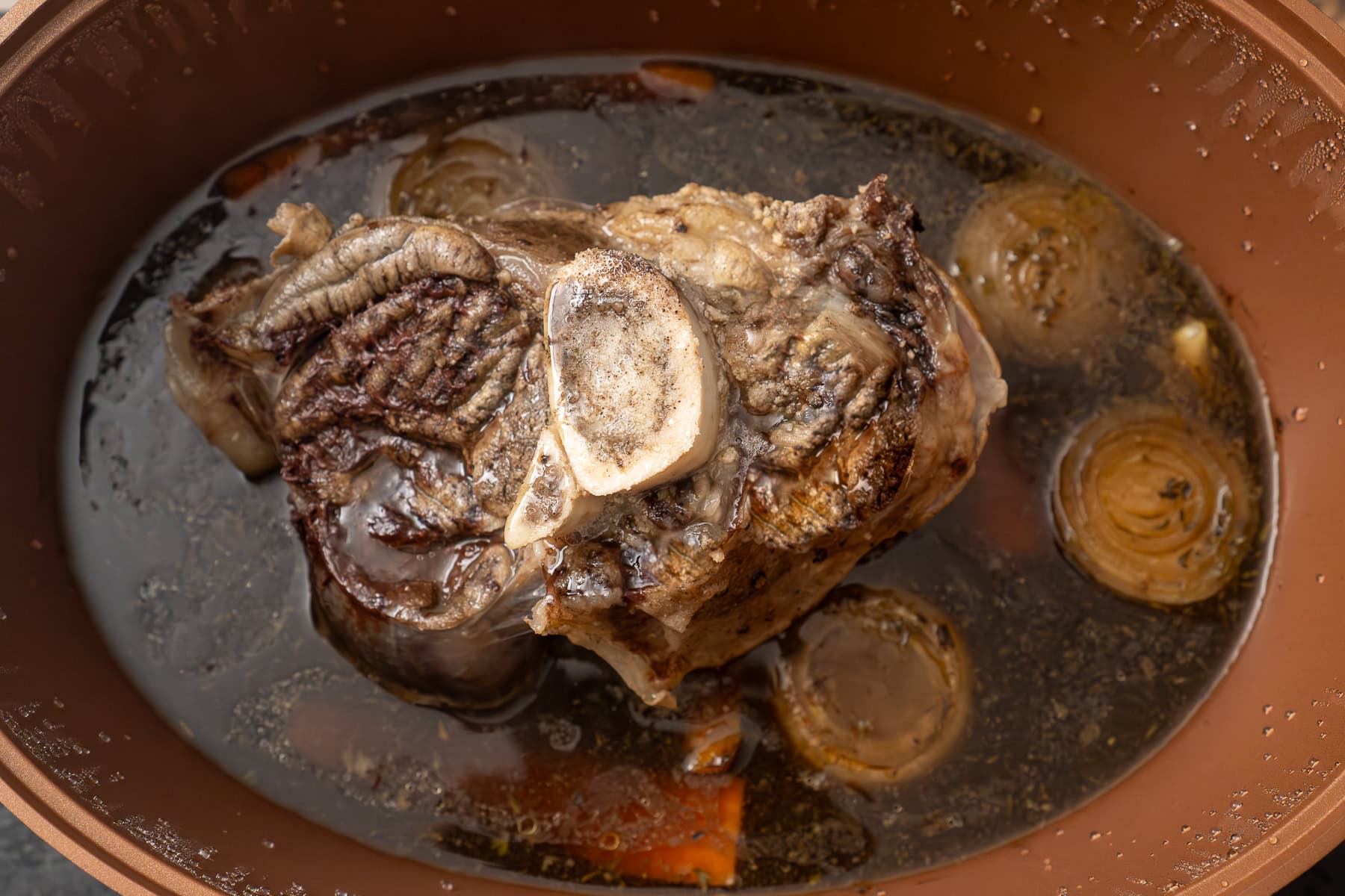 A cooked beef shank in a slow cooker sitting on top of beef stock and carrots and onions and garlic cloves