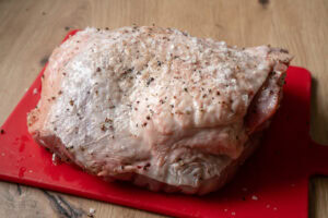 A turkey thigh coated in salt and pepper on a red chopping board
