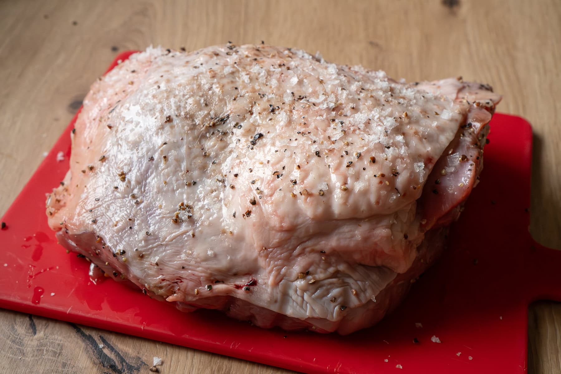 A turkey thigh coated in salt and pepper on a red chopping board