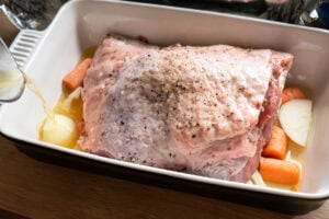 A turkey thigh coated in salt and pepper on a vegetable trivet in a oven proof dish with chicken stock being poured around the turkey thigh