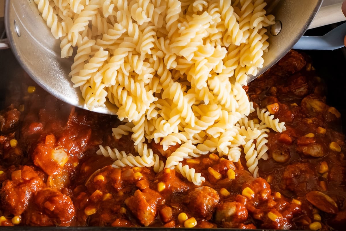 Pouring cooked pasta into sausage pasta bake tomato sauce