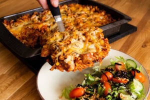 Putting sausage pasta bake with melted cheese on top onto a plate with salad on the side coated in balsamic vinegar